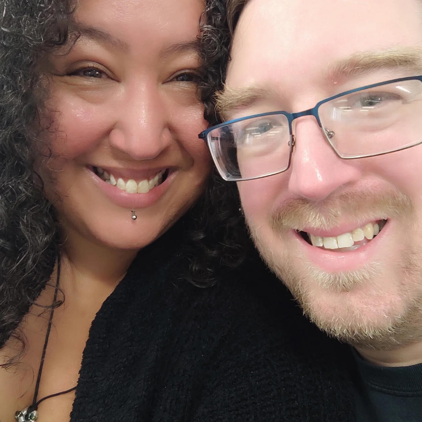 A photo of a mixed-ethnicity Black femme with curly hair and a white English man with glasses and a short beard, both smiling.